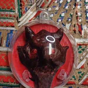 May include: A round, clear, plastic ornament with a red background. Inside the ornament is a dark brown, resin sculpture of a demon's head with a halo. The demon's head has a red, resin eye and two small, white, resin horns.