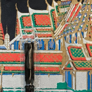 Könnte beinhalten: Ein schwarzer Amulet-Anhänger in einer transparenten Plastikverpackung. Der Anhänger ist in Form eines langen, dünnen Zylinders mit abgerundeter Oberseite. Die Verpackung befindet sich auf einem grün-weißen Hintergrund mit einem farbenfrohen Gemälde eines Tempels.