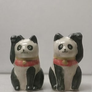 Puede incluir: Dos figuras de panda de cerámica con las patas levantadas. Los pandas tienen pelaje blanco y negro, collares rojos y pequeñas campanas doradas. Las figuras están sentadas erguidas, con una pata levantada en un gesto de llamada.