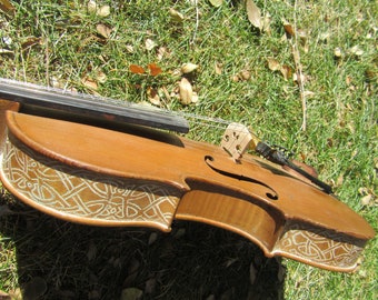 May include: A wooden violin with a Celtic knot design carved into the body. The violin is brown with a light brown finish and has a white string.