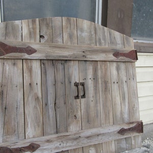 Könnte beinhalten: Eine verwitterte Holztür mit einem rustikalen Bogen oben und zwei eisernen Scharnieren. Die Tür ist aus wiederverwendeten Holzbrettern gefertigt und hat eine abgenutzte, graue Oberfläche. Die Tür hat einen Metallgriff mit dem Buchstaben "J" darauf.