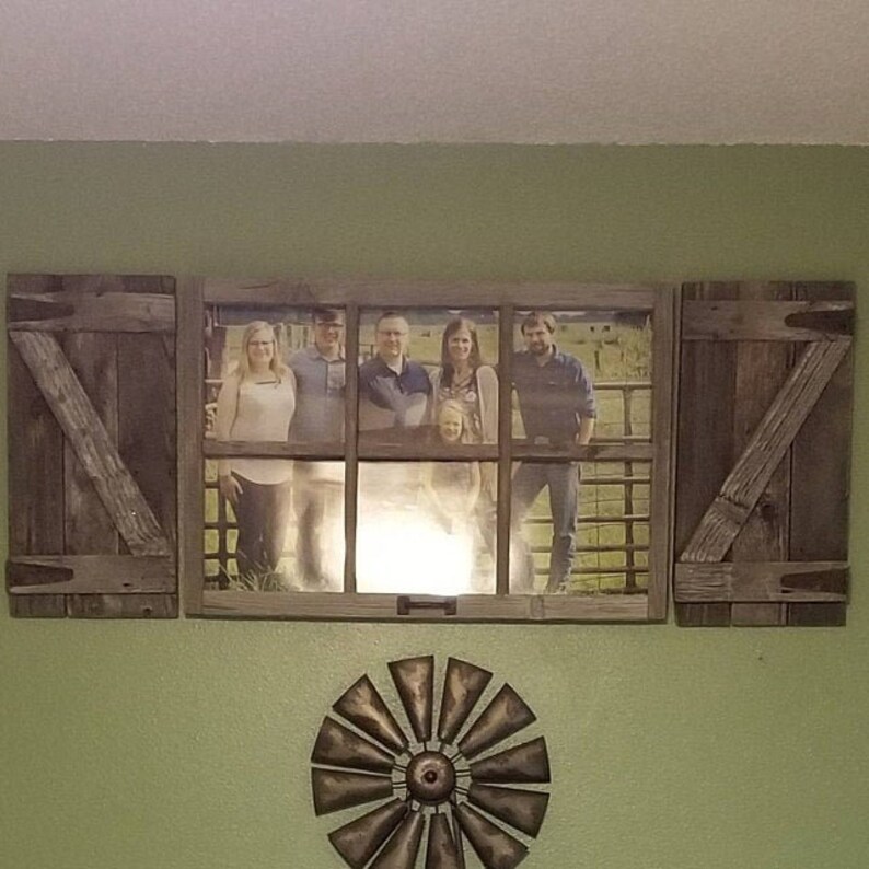 May include: A rustic wooden framed family photo with shutter-style side panels. The photo shows a family standing in front of a fence. A metal windmill decoration is below the framed photo, all mounted on a green wall.