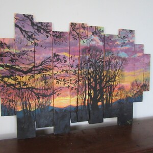 May include: A sunset scene painted on wooden planks, with a silhouette of trees and a purple, pink, and orange sky.