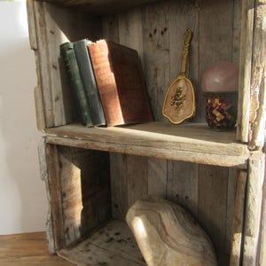 Könnte beinhalten: Rustikales Holzregal mit zwei gestapelten Kisten. Das obere Regal zeigt antike Bücher, einen dekorativen Handspiegel und eine rosa Kugel in einem Mosaikbecher. Das untere Regal enthält eine große Holzskulptur.