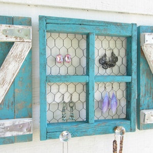 Könnte beinhalten: Ein türkisfarbener Holzrahmen mit einem Drahtgitter-Einsatz, der in vier Abschnitte unterteilt ist, um Ohrringe und Halsketten zu präsentieren. Der Rahmen hat eine abgenutzte weiße Lackierung und ist an einer weißen Wand montiert.