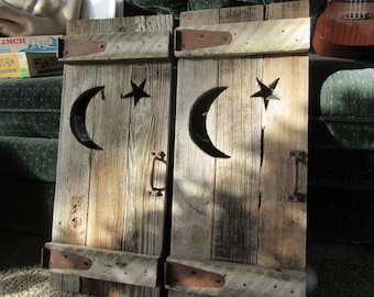 May include: Two rustic wooden shutters with cutouts of a crescent moon and a star. The shutters are painted a light brown color and have metal hinges and handles.