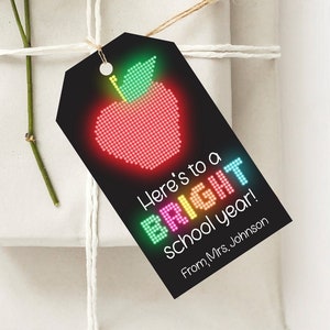 Puede incluir: Regalos envueltos atados con hilo y una etiqueta. La etiqueta negra presenta una manzana roja pixelada con un tallo verde y el texto "Here's to a BRIGHT school year!" y "From, Mrs. Johnson."