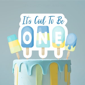 May include: A blue iced cake with blue, yellow, and green drips, decorated with a cake topper saying "It's Cool To Be ONE" and popsicle designs. The cake is on a white stand, set against a light blue backdrop.