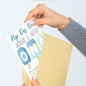 May include: A white card with blue text that reads "Pop On One Jackson is turning One". The card features a graphic of three blue and white popsicles with the letters "O N E" on them. The card is being held in a brown envelope.