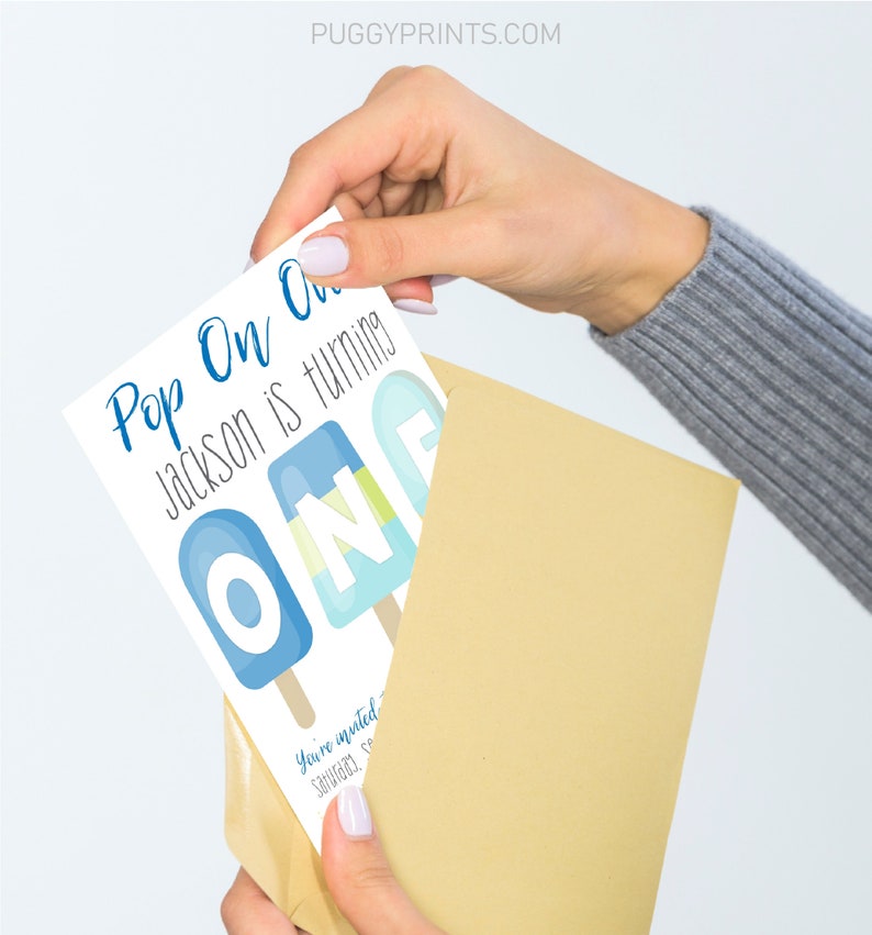 May include: A white card with blue text that reads "Pop On One Jackson is turning One". The card features a graphic of three blue and white popsicles with the letters "O N E" on them. The card is being held in a brown envelope.