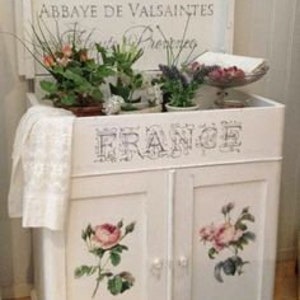 Puede incluir: Un mueble blanco con una parte superior enmarcada, con el texto "Le jardin des roses" y "Abbaye de Valsaintes". El mueble tiene dos puertas con ilustraciones de rosas y la palabra "FRANCE". Est&aacute; decorado con flores y un pa&ntilde;o blanco.