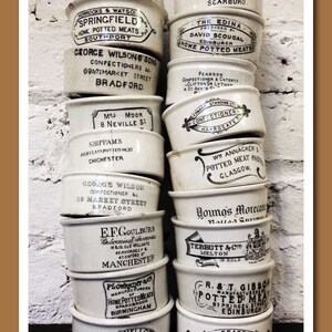 K&ouml;nnte beinhalten: Ein Stapel antiker, wei&szlig;er Keramik-Lebensmittelbeh&auml;lter mit schwarzem Text und Designs. Die runden Beh&auml;lter mit Deckeln zeigen verschiedene Firmennamen und Orte, wie "Manchester" und "Ipswich".
