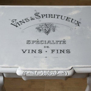 May include: A small, light gray table with ornate legs and a rectangular top. The top features black text that reads "Vins & Spiritueux" and "Sp&eacute;cialit&eacute; de Vins Fins". The table has a distressed, vintage appearance.