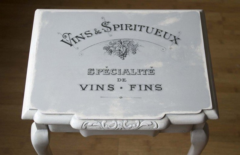 May include: A small, light gray table with ornate legs and a rectangular top. The top features black text that reads "Vins & Spiritueux" and "Sp&eacute;cialit&eacute; de Vins Fins". The table has a distressed, vintage appearance.
