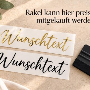 May include: Two rectangular adhesive labels with the German word "Wunschtext" in a cursive script font. One label features metallic gold lettering, while the other has black lettering. A black plastic squeegee tool is positioned to the right.