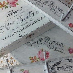 Puede incluir: Perchero de madera blanca envejecida con detalles florales y escritura francesa. Presenta rosas rosas y ganchos de estilo vintage. El perchero tiene el texto "Fleur Mme Boileve" y "Maison C. Riffard".
