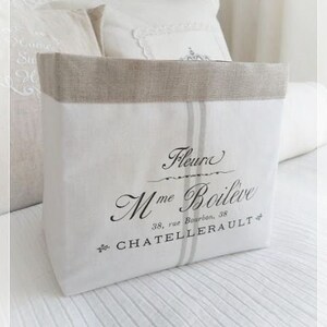 Puede incluir: Una bolsa de almacenamiento de lino blanco con un borde de lino beige. La bolsa presenta el texto "Fleur Mme Boil&egrave;ve" y "38, rue Bourbon, CHATELLERAULT" en escritura negra. Dos rayas verticales grises recorren la parte delantera.