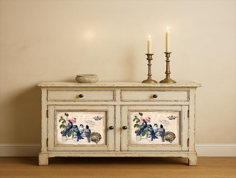 Puede incluir: Un aparador blanquecino envejecido con dos cajones y dos puertas. Cada puerta presenta una imagen enmarcada de p&aacute;jaros azules, flores rosas y texto. Dos candelabros de lat&oacute;n con velas encendidas se encuentran encima, junto con un peque&ntilde;o cuenco tejido.