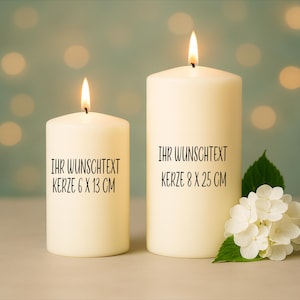 May include: Two ivory pillar candles with lit flames. The larger candle is 8 cm x 25 cm, and the smaller is 6 cm x 13 cm. Text on the candles reads "IHR WUNSCHTEXT". A sprig of white flowers and green leaves is on the right.