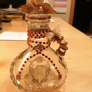 K&ouml;nnte beinhalten: Eine dekorative Glasflasche mit einem cherubf&ouml;rmigen Verschluss und einem bienenf&ouml;rmigen Griff. Die Flasche ist mit roten und goldenen Verzierungen, darunter Strasssteine und Blumenakzente, verziert. Die Worte "Home sweet Home" sind aufgedruckt.