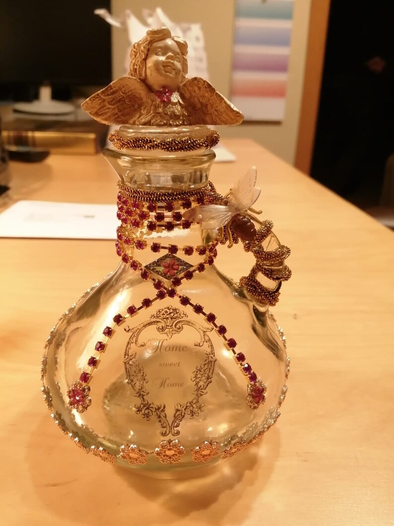 K&ouml;nnte beinhalten: Eine dekorative Glasflasche mit einem cherubf&ouml;rmigen Verschluss und einem bienenf&ouml;rmigen Griff. Die Flasche ist mit roten und goldenen Verzierungen, darunter Strasssteine und Blumenakzente, verziert. Die Worte "Home sweet Home" sind aufgedruckt.