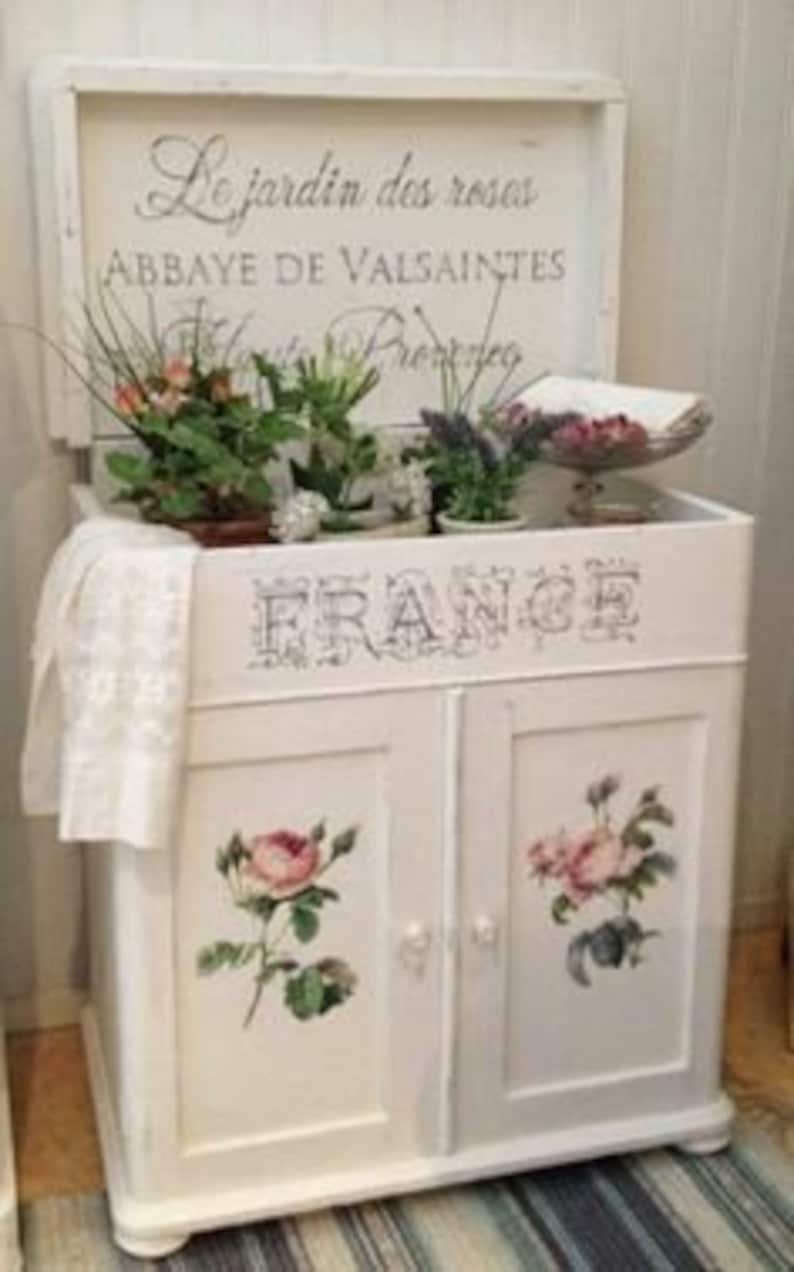 Puede incluir: Un mueble blanco con acabado envejecido, con ilustraciones florales y la palabra "FRANCE" estampada en la parte delantera. La secci&oacute;n superior muestra el texto "Le jardin des roses" y "ABBAYE DE VALSAINTES". Un pa&ntilde;o de encaje blanco y arreglos florales adornan la parte superior.