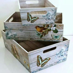 Puede incluir: Tres cajas de almacenamiento de madera apiladas, cada una con un recorte de asa. Las cajas est&aacute;n pintadas con un efecto de veta de madera envejecida y decoradas con mariposas, p&aacute;jaros y dise&ntilde;os florales en tonos azules, marrones y blancos. Las cajas est&aacute;n dise&ntilde;adas para almacenamiento decorativo.