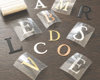 May include: A collection of adhesive vinyl letters in various colors including gold, copper, white, and black, featuring different serif and sans-serif fonts. A white plastic application squeegee with a felt edge is positioned in the upper left corner.