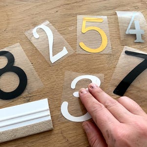 Könnte beinhalten: Eine Sammlung von selbstklebenden Vinyl-Hausnummern mit den Ziffern 8, 2, 5, 4, 3 und 7 in den Farben Schwarz, Weiß und Gold, jeweils auf einer transparenten Trägerfolie, sowie einem weißen Kunststoff-Rakel mit Filzkante zum Auftragen.