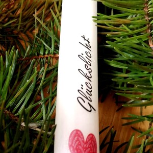 May include: A white candle with the word "Gl&uuml;ckslicht" and a red heart design. The candle is surrounded by green pine needles, creating a festive and natural aesthetic. Ideal for home decor or special occasions.