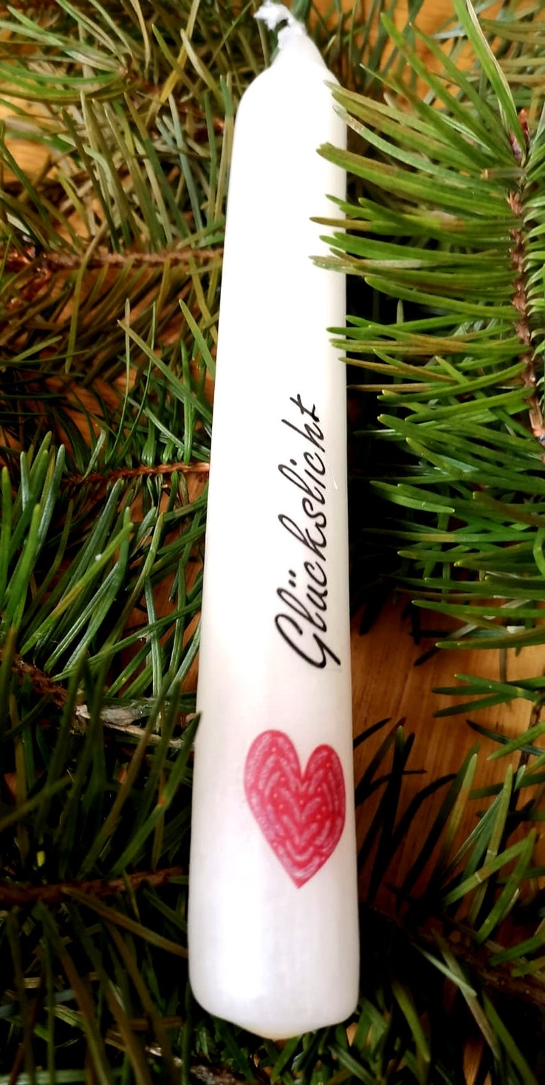 May include: A white candle with the word "Gl&uuml;ckslicht" and a red heart design. The candle is surrounded by green pine needles, creating a festive and natural aesthetic. Ideal for home decor or special occasions.