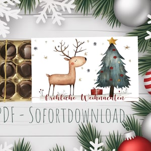 Könnte beinhalten: Eine Weihnachtskarte mit einer Illustration eines Rentiers und eines geschmückten Weihnachtsbaums. Die Karte trägt den Text "Fröhliche Weihnachten" und ist von Weihnachtsschmuck umgeben. Links ist eine Schachtel Pralinen zu sehen.
