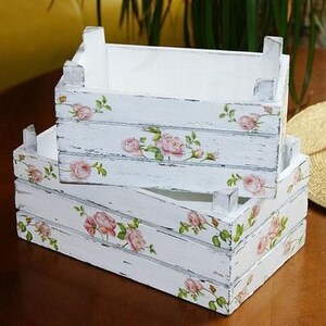 Puede incluir: Dos cajas de madera blancas apiladas, con un acabado envejecido. Cada caja est&aacute; decorada con patrones florales de rosas rosas y hojas verdes. Las cajas est&aacute;n sobre una superficie de madera marr&oacute;n oscuro.