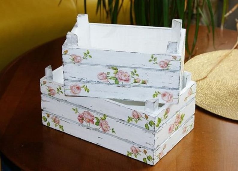 Puede incluir: Dos cajas de madera blancas apiladas, con un acabado envejecido. Cada caja est&aacute; decorada con patrones florales de rosas rosas y hojas verdes. Las cajas est&aacute;n sobre una superficie de madera marr&oacute;n oscuro.