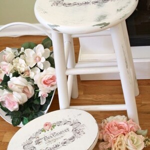 Puede incluir: Dos taburetes de madera blanca envejecida con asientos redondos. Los asientos presentan un dise&ntilde;o floral y el texto "Paul Charvet". Un ramo de rosas rosas y blancas y otras flores se encuentra cerca, a&ntilde;adiendo un toque de elegancia a la escena.