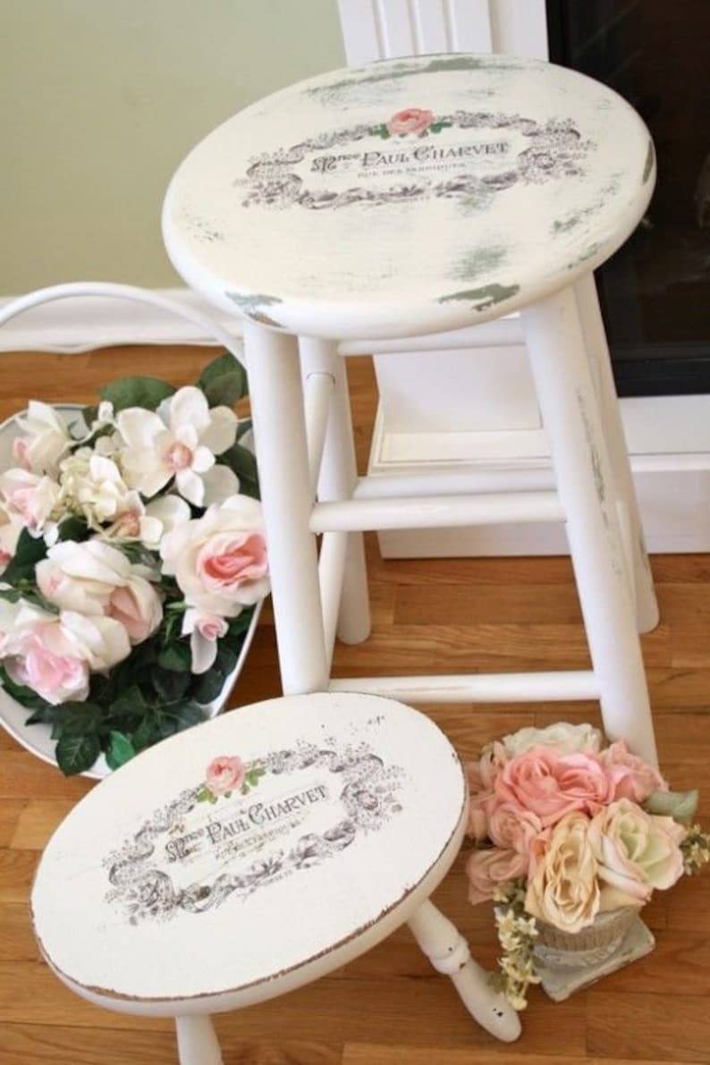 Puede incluir: Dos taburetes de madera blanca envejecida con asientos redondos. Los asientos presentan un dise&ntilde;o floral y el texto "Paul Charvet". Un ramo de rosas rosas y blancas y otras flores se encuentra cerca, a&ntilde;adiendo un toque de elegancia a la escena.