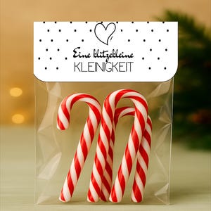 May include: A transparent cellophane bag holding four red and white striped candy canes. The bag features a white label with black polka dots, a heart, and the text "Eine klitzekleine KLEINIGKEIT".