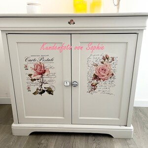 Puede incluir: Un mueble blanco con dos puertas, adornado con calcoman&iacute;as florales y escritura. Cada puerta presenta un dise&ntilde;o de rosa diferente, con un pomo de cristal. El mueble tiene una parte superior y una base decorativas.