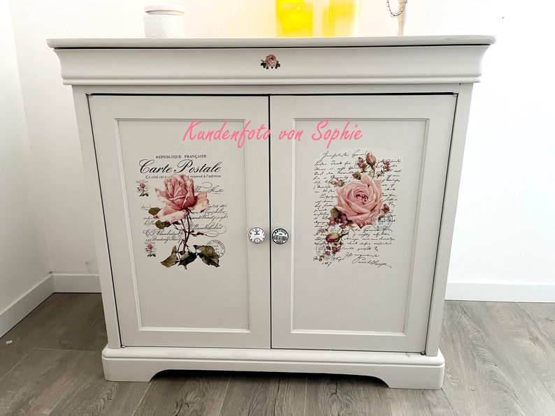 Puede incluir: Un mueble blanco con dos puertas, adornado con calcoman&iacute;as florales y escritura. Cada puerta presenta un dise&ntilde;o de rosa diferente, con un pomo de cristal. El mueble tiene una parte superior y una base decorativas.