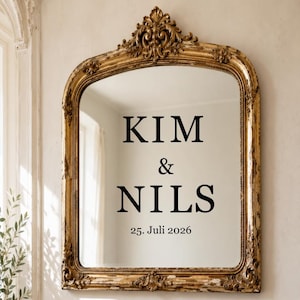 May include: An arched wall mirror with an ornate, distressed gold frame featuring intricate scrollwork at the top. The glass surface displays black text reading KIM & NILS followed by the date 25. Juli 2026 centered in the middle of the mirror.