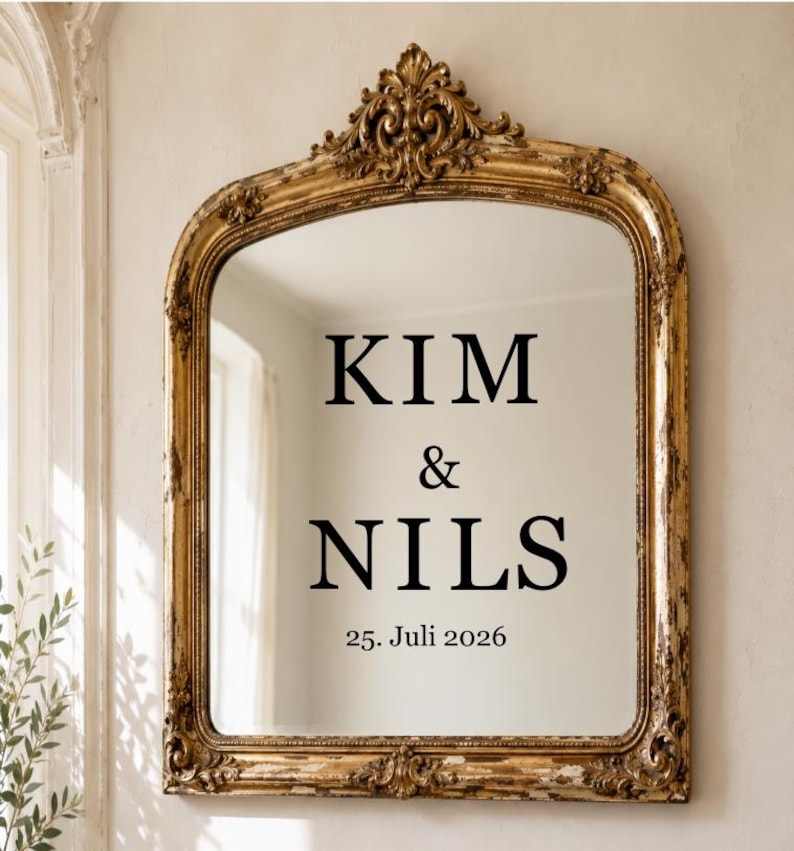 May include: An arched wall mirror with an ornate, distressed gold frame featuring intricate scrollwork at the top. The glass surface displays black text reading KIM & NILS followed by the date 25. Juli 2026 centered in the middle of the mirror.