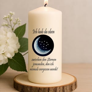 Può includere: Candela a colonna color avorio con fiamma accesa, con un design a mezzaluna e stelle. La candela poggia su una fetta di legno e presenta testo in tedesco. Sullo sfondo un'arrangiamento floreale bianco.