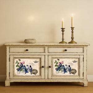 Puede incluir: Un aparador blanquecino envejecido con dos cajones y dos puertas. Cada puerta presenta una imagen enmarcada de p&aacute;jaros azules, flores rosas y texto. Dos candelabros de lat&oacute;n con velas encendidas se encuentran encima, junto con un peque&ntilde;o cuenco tejido.