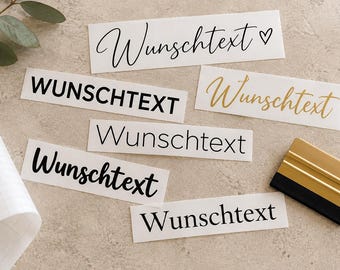 May include: Six rectangular adhesive vinyl decals featuring the German word Wunschtext in various fonts and colors, including black, gold, and script styles, accompanied by a roll of transfer paper and a gold and black plastic squeegee tool.