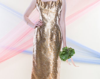 May include: A woman in a gold brocade dress holds a bouquet of green succulents. She is standing in front of a backdrop of pink and blue fabric.