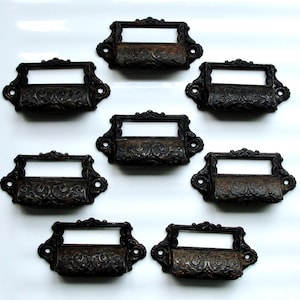 Puede incluir: Ocho tiradores de cajón de metal negro antiguo con diseños ornamentados y un portaetiquetas rectangular.