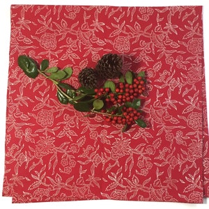 May include: A red tablecloth with a white floral pattern. The tablecloth is decorated with a sprig of green leaves, red berries, and two pinecones. The tablecloth is square and folded.