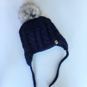 Puede incluir: Un gorro de punto azul marino con un pompón de piel sintética blanca y lazos largos. El gorro tiene una etiqueta tejida que dice "RD".