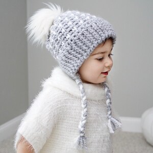 Op de afbeelding: Een grijze en witte gebreide baby muts met een witte pompon en witte gevlochten banden. De muts wordt gedragen door een kind.
