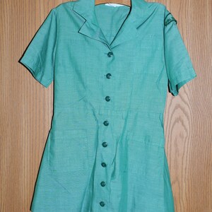 May include: A vintage green Girl Guide uniform dress with short sleeves and a button-up front. The dress has two pockets and a patch on the right sleeve.